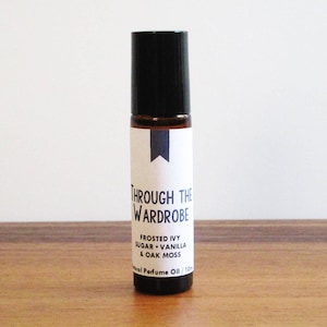 May include: A small brown glass bottle with a black rollerball top. The bottle has a white label with the text "Through the Wardrobe" and "Frosted Ivy, Sugar, Vanilla & Oak Moss" in black text. The bottle is sitting on a wooden surface.