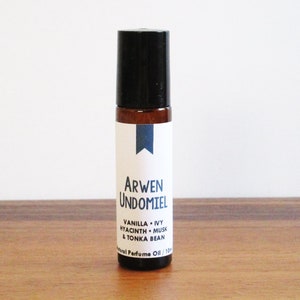 May include: A brown glass roll-on bottle with a black cap and a white label. The label reads "Arwen Undomiel" and lists the scent notes: "Vanilla + Ivy, Hyacinth + Musk & Tonka Bean". The bottle is labeled "Natural Perfume Oil 13ml".