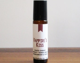 May include: A brown glass roll-on bottle with a black cap and a white label. The label says "Vampire's Kiss" and lists the scent notes: "Black Cherry, Violet, Tonka Bean, Sandalwood & Musk". The bottle is labeled "10ml Perfume Oil".