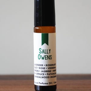 May include: A small, brown glass bottle with a black cap. The label reads "SALLY OWENS" with a list of ingredients. The bottle is on a wooden surface. The product is a natural perfume oil.