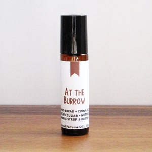 May include: A brown glass roll-on bottle with a black cap and a white label that reads "AT THE BURROW" and lists the ingredients "BREAD - CINNAMON - BROWN SUGAR - NUTMEG - MAPLE SYRUP & BUTTER" and "Natural Perfume Oil / 10 ml".