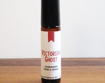 May include: A small brown glass bottle with a black rollerball top. The bottle has a white label with the text "Victorian Ghost" in red lettering. The label also includes the text "Cardamom Rose & Cedar" and "Natural Perfume Oil 1/3 oz."