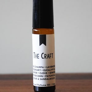 May include: A small, brown glass bottle with a black cap. The bottle has a white label with the text "THE CRAFT" and a list of ingredients. The bottle is sitting on a wooden surface.