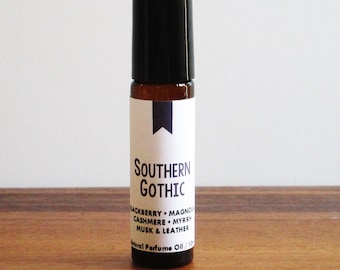 May include: A brown glass roll-on bottle with a black cap and a white label that reads "Southern Gothic" and lists the fragrance notes: "Blackberry, Magnolia, Cashmere, Myrrh, Musk & Leather". Natural Perfume Oil.