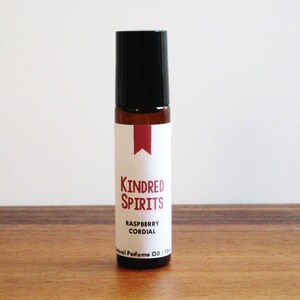 May include: A small brown glass bottle with a black rollerball top. The bottle has a white label with red text that reads "KINDRED SPIRITS RASPBERRY CORDIAL Natural Perfume Oil 10ml".