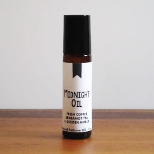 May include: A brown glass roll-on bottle with a black cap and a white label that reads "MIDNIGHT OIL" and "FRESH COFFEE BERGAMOT TEA & GOLDEN AMBER" with the text "Natural Perfume Oil / 10 ml" at the bottom.