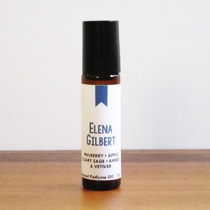 May include: A brown glass roller bottle with a black cap and a white label. The label says "Elena Gilbert" and lists the scent as "Mulberry + Apple, Clary Sage + Amber & Vetiver". The label also says "Natural Perfume Oil 1/3 oz."