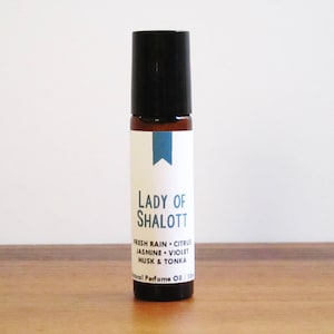 May include: A brown glass roller bottle with a black cap and a white label. The label says "Lady of Shalott" and lists the fragrance notes: "Fresh Rain, Citrus, Jasmine, Violet, Musk & Tonka". The label also says "Natural Perfume Oil / 10 ml".
