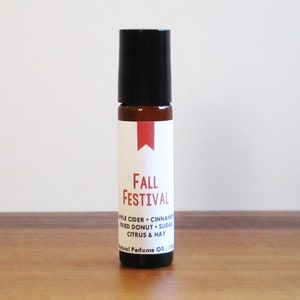 May include: A small, brown glass roll-on bottle with a black cap. The label reads "Fall Festival" with a red banner and lists scent notes: apple cider, cinnamon, fried donut, sugar, citrus & hay. The bottle is on a wooden surface.