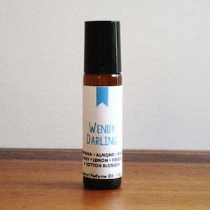 May include: A brown glass roller bottle with a black cap and a white label. The label says "WENDY DARLING" and lists the ingredients: "Vanilla, Almond, Coconut, Lemon, Freesia & Cotton Blossom". The label also says "Natural Perfume Oil 1/10 oz."