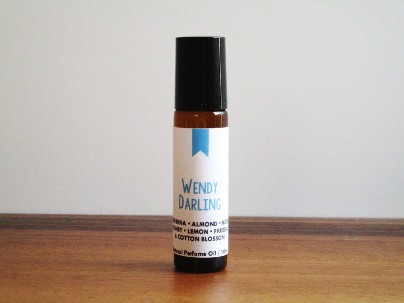 May include: A brown glass roller bottle with a black cap and a white label. The label says "WENDY DARLING" and lists the ingredients: "Vanilla, Almond, Coconut, Lemon, Freesia & Cotton Blossom". The label also says "Natural Perfume Oil 1/10 oz."