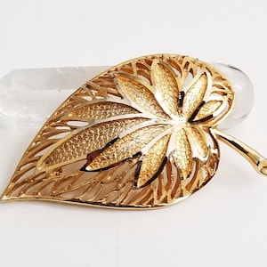 May include: A gold-tone leaf-shaped brooch with a detailed, textured design. The leaf is openwork and has a delicate, intricate pattern.