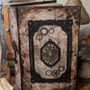 Könnte beinhalten: Ein Notizbuch im Steampunk-Stil mit einem Kartenmotiv auf dem Einband. Das Notizbuch hat einen schwarzen Zierrahmen mit Zahnrädern und verzierten Eckakzenten. Der Einband hat einen verwitterten, Vintage-Look mit messingfarbenen Nieten und einem Verschluss.