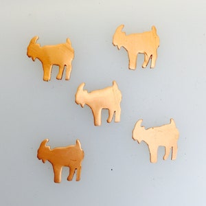 Puede incluir: Cinco pequeños dijes con forma de cabra de color cobre están dispersos sobre una superficie gris claro. Cada dije tiene una silueta detallada de una cabra con cuernos, barba y cuatro patas. Los dijes son probablemente para hacer joyas u otras manualidades.
