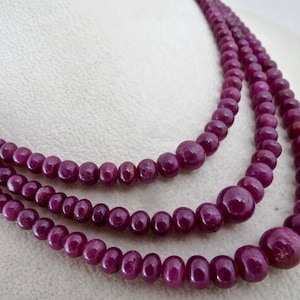 May include: Three strands of a ruby bead necklace. The beads are a deep, rich red-purple color. The necklace is displayed on a white surface, showcasing the jewelry's elegant design. The beads vary in size, creating a layered effect.
