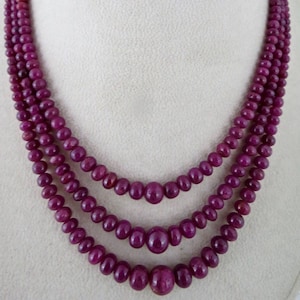 May include: A three-strand necklace featuring polished, round, deep red beads. The necklace is displayed on a white mannequin, showcasing the graduated sizes of the beads, with larger beads at the center of each strand.
