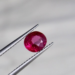 May include: A vibrant red ruby gemstone held in tweezers. The ruby is round and has a smooth, polished surface.