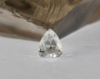 May include: A clear pear-shaped gemstone with facets, resting on a white surface.
