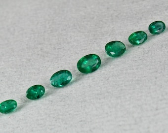 May include: Seven oval-shaped green emeralds of varying sizes arranged in a line on a white surface.