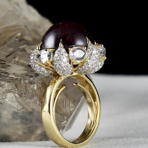 Könnte beinhalten: Ein goldener Ring mit einem großen, runden, dunkelroten Edelstein, der von kleineren, klaren Edelsteinen umgeben ist. Der Ring hat ein zartes, florales Design.