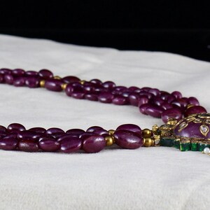 Pu&ograve; includere: Una collana multifilo realizzata con perline viola scuro con una chiusura dorata e un grande ciondolo con una pietra viola circondata da pietre pi&ugrave; piccole di colore verde e bianco.