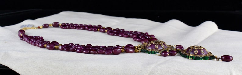 Pu&ograve; includere: Una collana multifilo realizzata con perline viola scuro con una chiusura dorata e un grande ciondolo con una pietra viola circondata da pietre pi&ugrave; piccole di colore verde e bianco.