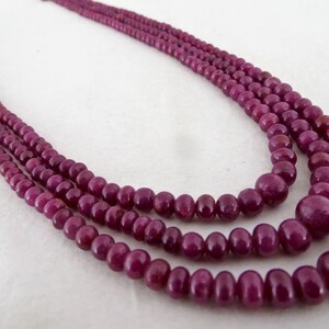 May include: A three-strand necklace featuring polished, deep red-purple beads. The beads vary in size, with larger ones at the front and smaller ones towards the back. The necklace is likely made of gemstones and is displayed against a neutral background.