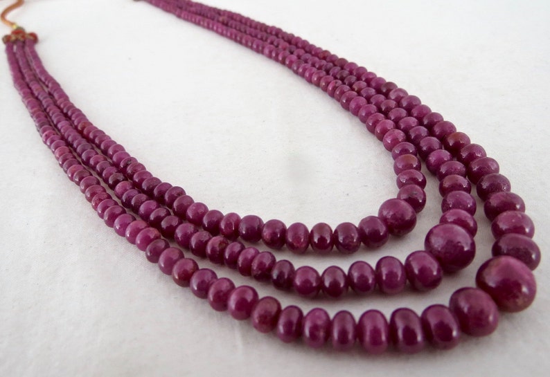 May include: A three-strand necklace featuring polished, deep red-purple beads. The beads vary in size, with larger ones at the front and smaller ones towards the back. The necklace is likely made of gemstones and is displayed against a neutral background.
