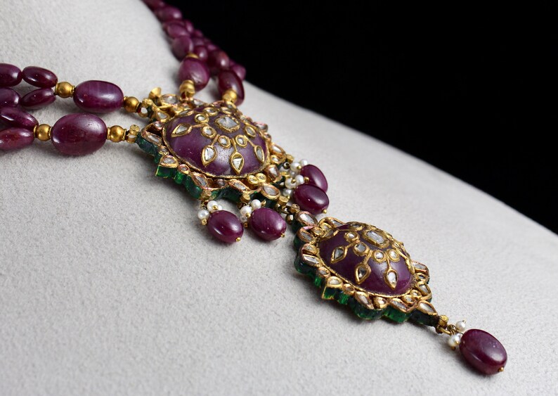 Pu&ograve; includere: Una collana d'oro con un ciondolo di rubino e diamanti. Il ciondolo &egrave; composto da due rubini rotondi circondati da diamanti pi&ugrave; piccoli e pietre verdi.