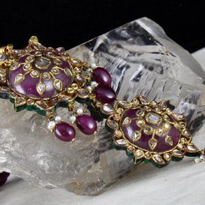 Pu&ograve; includere: Una collana d'oro con due grandi pendenti che presentano una pietra viola circondata da diamanti ed emeraldi pi&ugrave; piccoli. La collana &egrave; realizzata con perle di rubino.