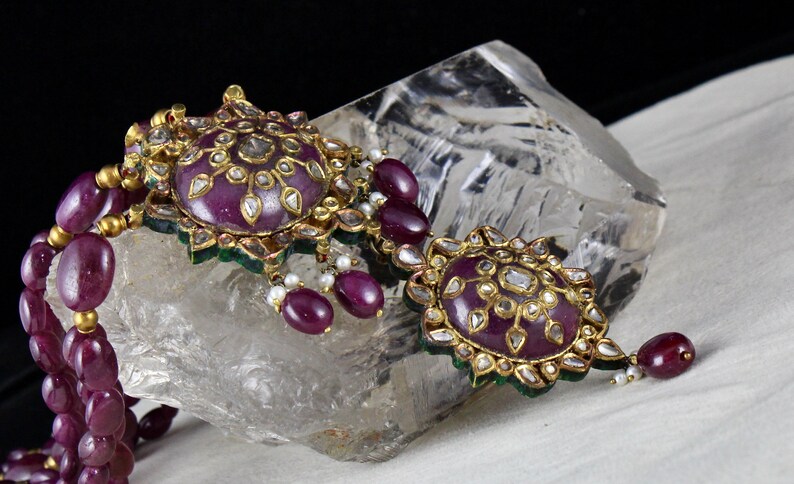 Pu&ograve; includere: Una collana d'oro con due grandi pendenti che presentano una pietra viola circondata da diamanti ed emeraldi pi&ugrave; piccoli. La collana &egrave; realizzata con perle di rubino.