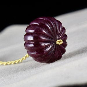 May include: A dark purple, carved gemstone bead with a gold accent. The bead is attached to a gold cord.