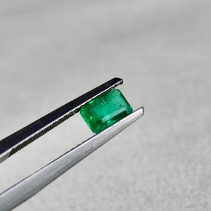 May include: A vibrant green emerald gemstone held in tweezers. The emerald is cut into a rectangular shape with facets.