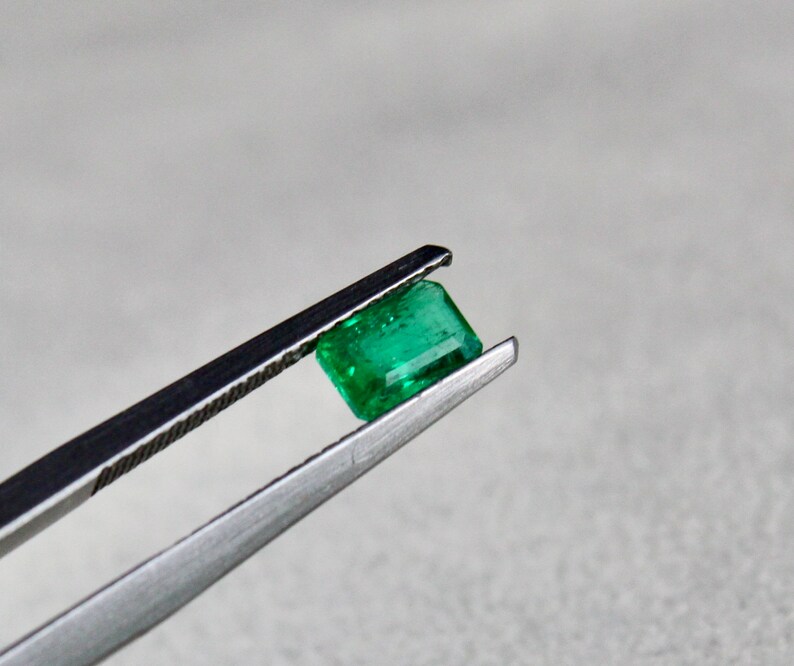 May include: A vibrant green emerald gemstone held in tweezers. The emerald is cut into a rectangular shape with facets.