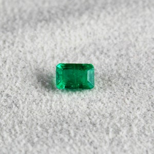 May include: A vibrant green emerald gemstone, cut into a rectangular shape with facets, is displayed on a textured gray surface.
