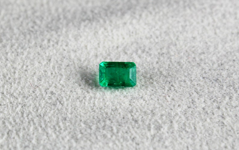May include: A vibrant green emerald gemstone, cut into a rectangular shape with facets, is displayed on a textured gray surface.