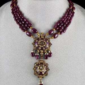 Pu&ograve; includere: Una collana d'oro con un ciondolo con un designo di rubino e diamanti. La collana &egrave; composta da tre fili di perle di rubino.