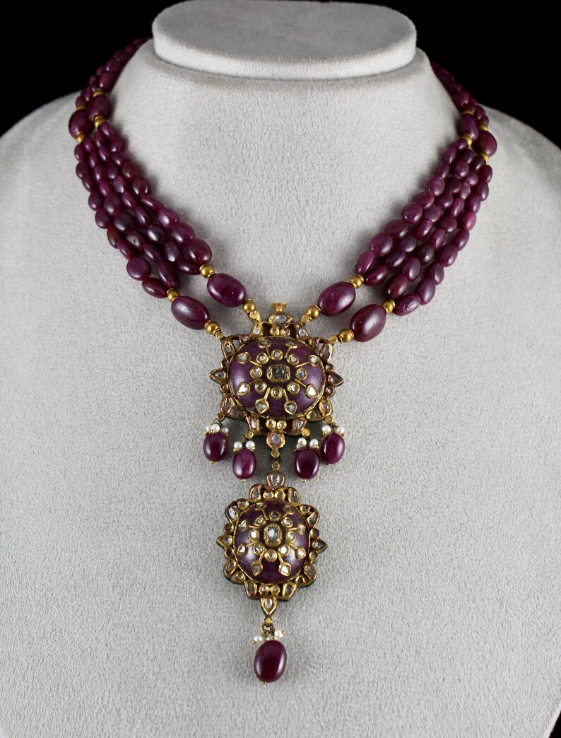 Pu&ograve; includere: Una collana d'oro con un ciondolo con un designo di rubino e diamanti. La collana &egrave; composta da tre fili di perle di rubino.