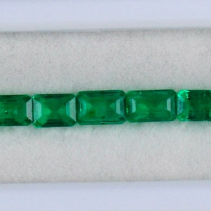 May include: A row of 17 emerald green rectangular gemstones in a white display case.