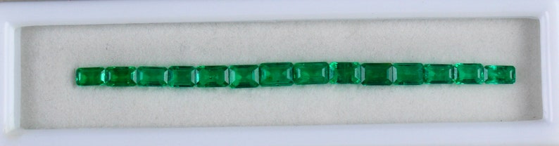 May include: A row of 17 emerald green rectangular gemstones in a white display case.