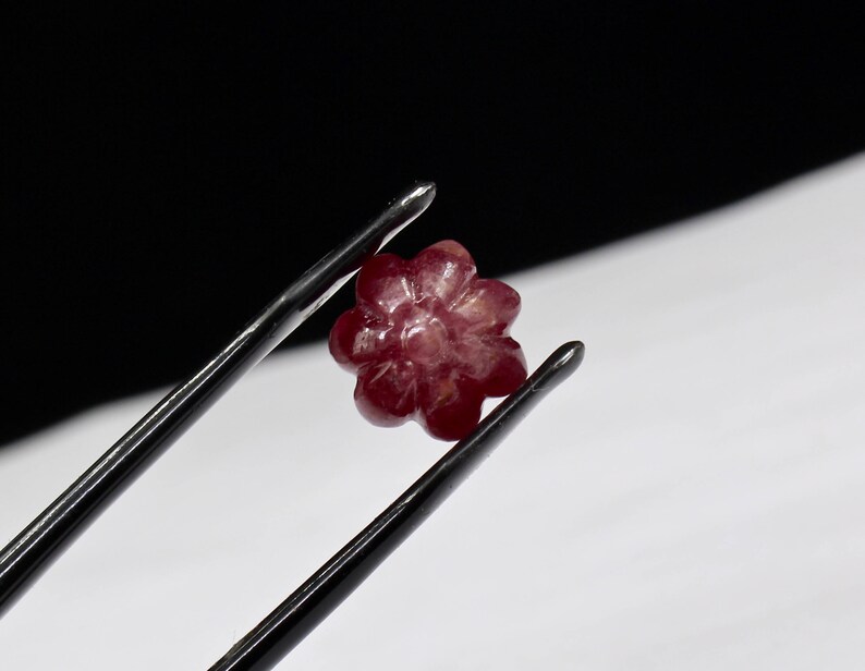 May include: A carved ruby gemstone in the shape of a flower. The gemstone is held by a pair of tweezers.