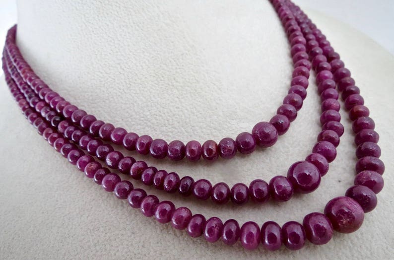 May include: Three strands of a ruby bead necklace. The beads are a deep, rich red-purple color. The necklace is displayed on a white surface, showcasing the jewelry's elegant design. The beads vary in size, creating a layered effect.