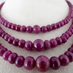 May include: Three strands of ruby bead necklaces. The beads are a deep, rich red-purple color. The necklaces are displayed on a white surface, showcasing the jewelry's color and design. The beads vary in size.