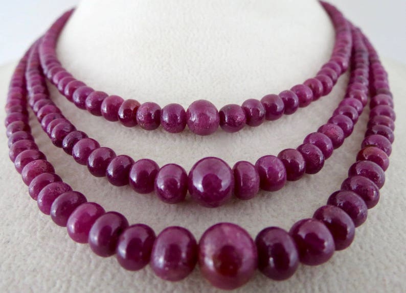 May include: Three strands of ruby bead necklaces. The beads are a deep, rich red-purple color. The necklaces are displayed on a white surface, showcasing the jewelry's color and design. The beads vary in size.