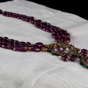 Pu&ograve; includere: Una collana a tre file con un grande ciondolo decorato. La collana &egrave; fatta di perline viola scuro e il ciondolo &egrave; in oro con pietre verdi e bianche.