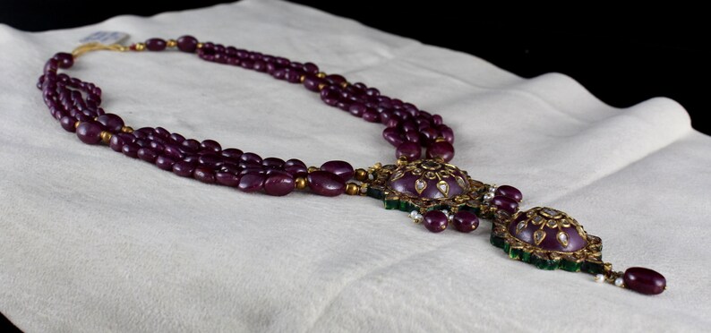 Pu&ograve; includere: Una collana a tre file con un grande ciondolo decorato. La collana &egrave; fatta di perline viola scuro e il ciondolo &egrave; in oro con pietre verdi e bianche.