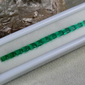 May include: A row of emerald green gemstones in a white rectangular box. The gemstones are cut into square shapes and are arranged in a line.
