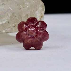 May include: A carved pink ruby flower gemstone. The flower has five petals and a center with a raised design.