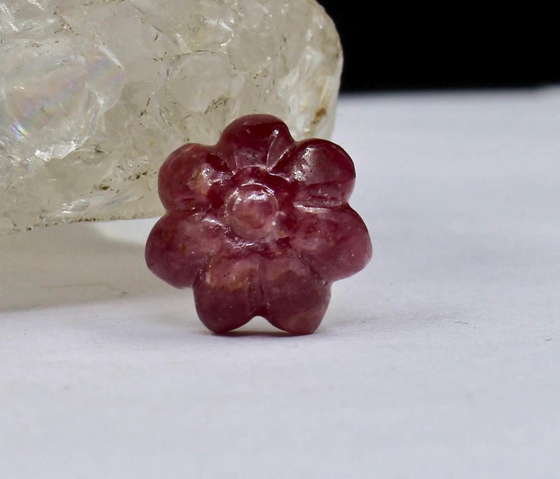 May include: A carved pink ruby flower gemstone. The flower has five petals and a center with a raised design.