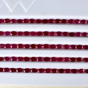 May include: A row of 20 oval-shaped, deep red gemstones arranged in five rows of four on a white surface.